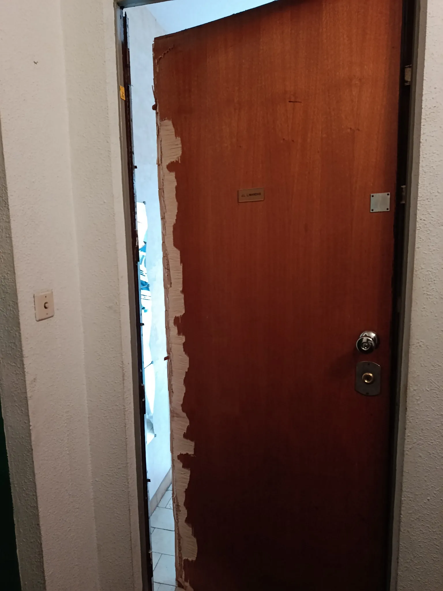 Porte intérieure en bois brun entrouverte, avec le bord du panneau fortement endommagé et une serrure visible.