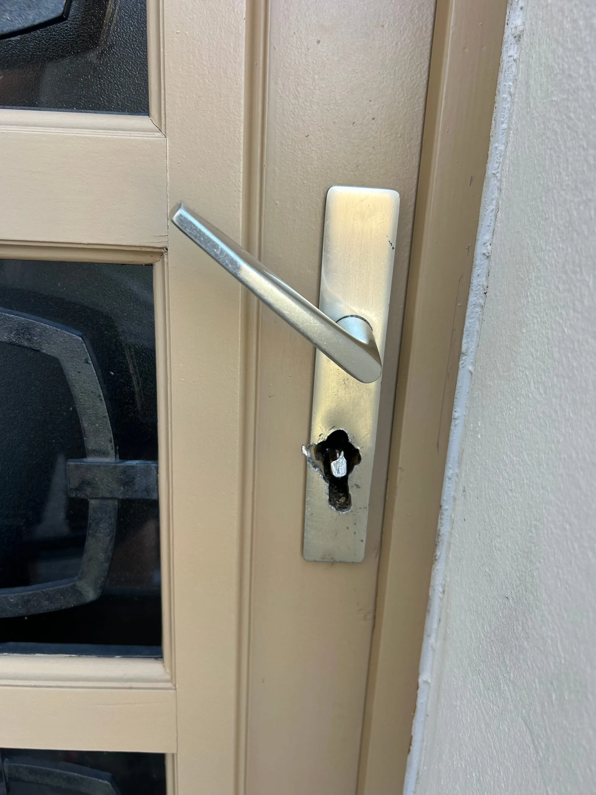 Plaque de poignée percée autour du logement du cylindre sur une porte d’entrée beige.