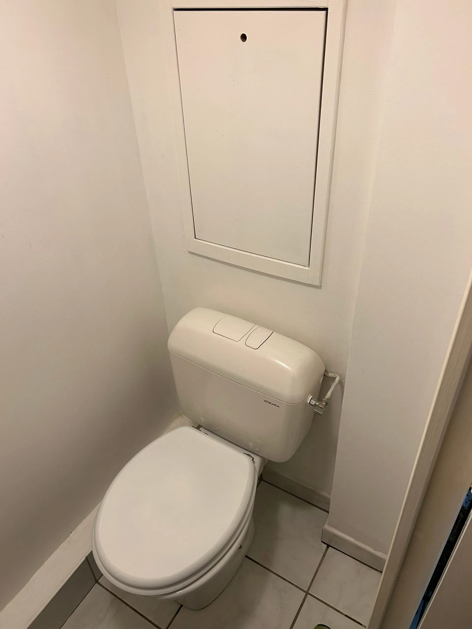 Ancien WC installé sous une trappe technique murale blanche.