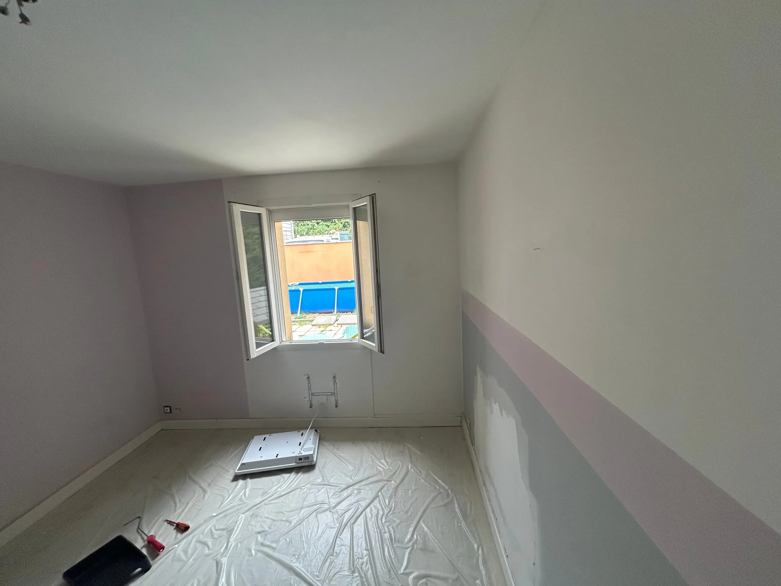 Pièce intérieure en cours de peinture, avec murs protégés, bâche au sol et fenêtre ouverte sur l’extérieur.