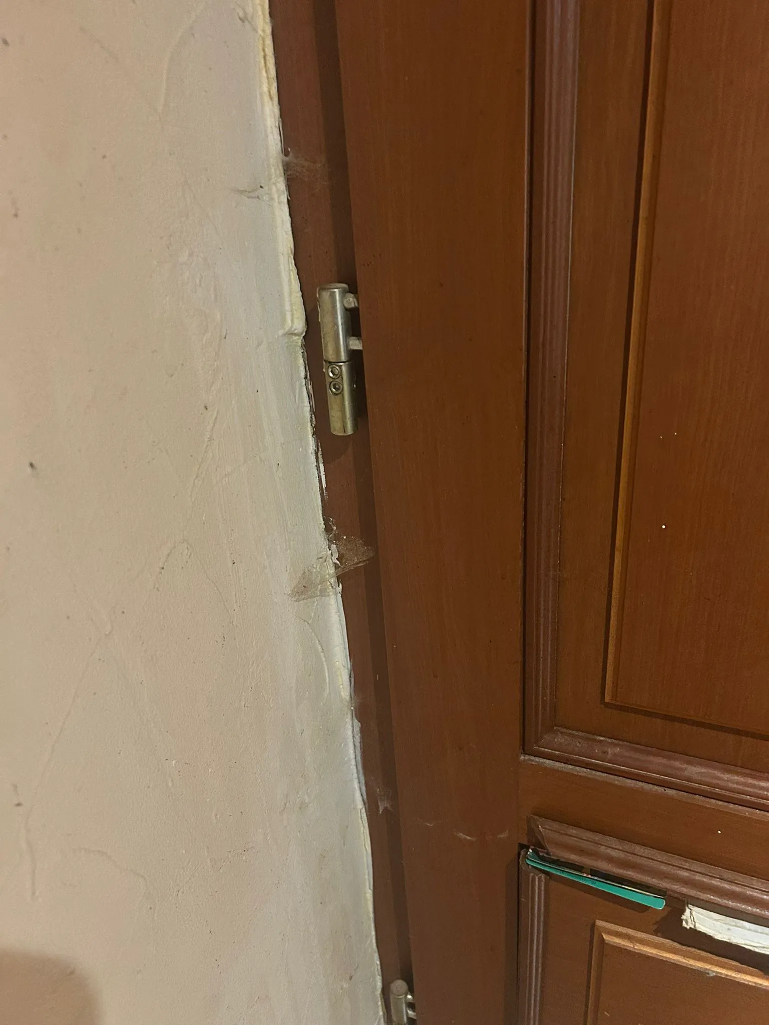 Charnière métallique sur une porte en bois brun près d’un mur clair.