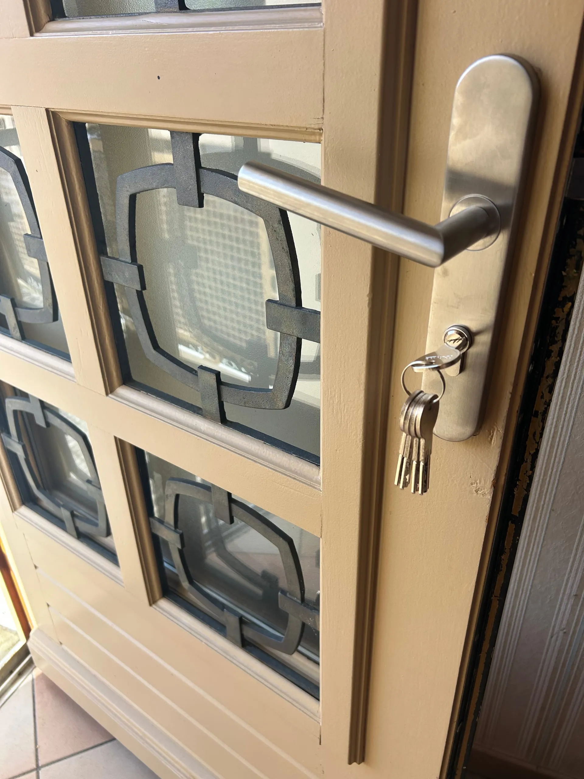 Cylindre européen neuf avec ses clés sous la poignée métallique d’une porte vitrée beige.