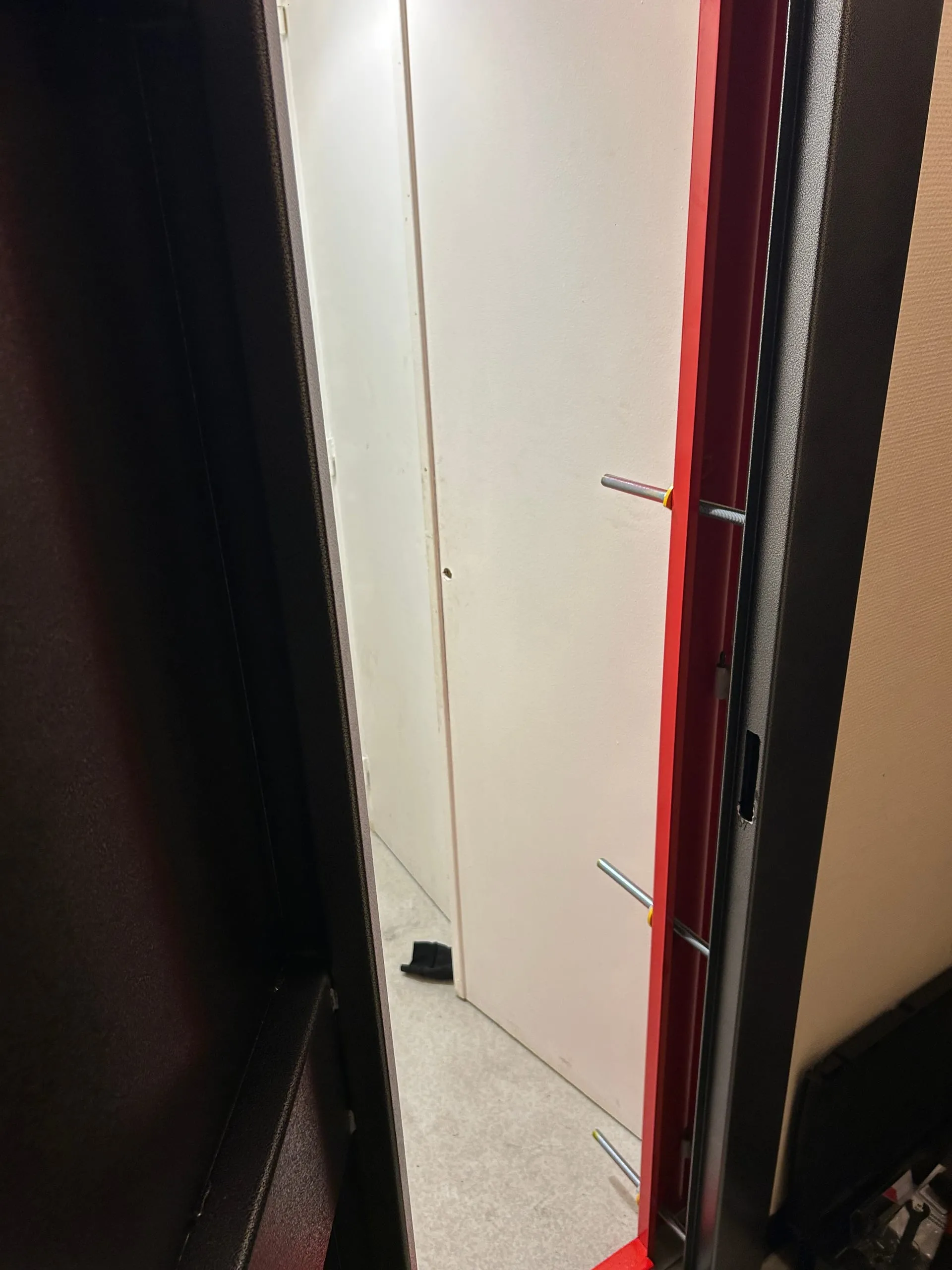 Porte anti-squat entrouverte laissant voir son encadrement rouge, ses tiges de scellement et le couloir intérieur.